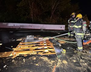 Two firefighters in full gear spray water on burnt wooden pallets and debris on a dark rooftop at night. Smoke rises from the charred remains, and trees are visible in the background.