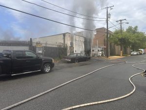 Smoke billows from a damaged industrial building as fire hoses stretch along the street. Two parked vehicles, including a black pickup and a dark sedan, are near the building. Firefighters and trees are visible in the background.