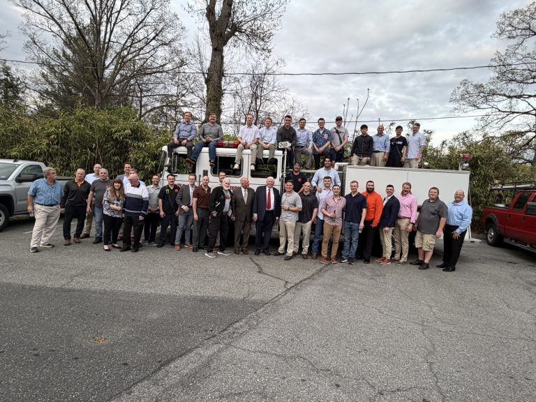 A large group of people, mostly men in business or casual attire, stand and sit in front of and on top of a fire truck in a parking lot, with trees and cars in the background.