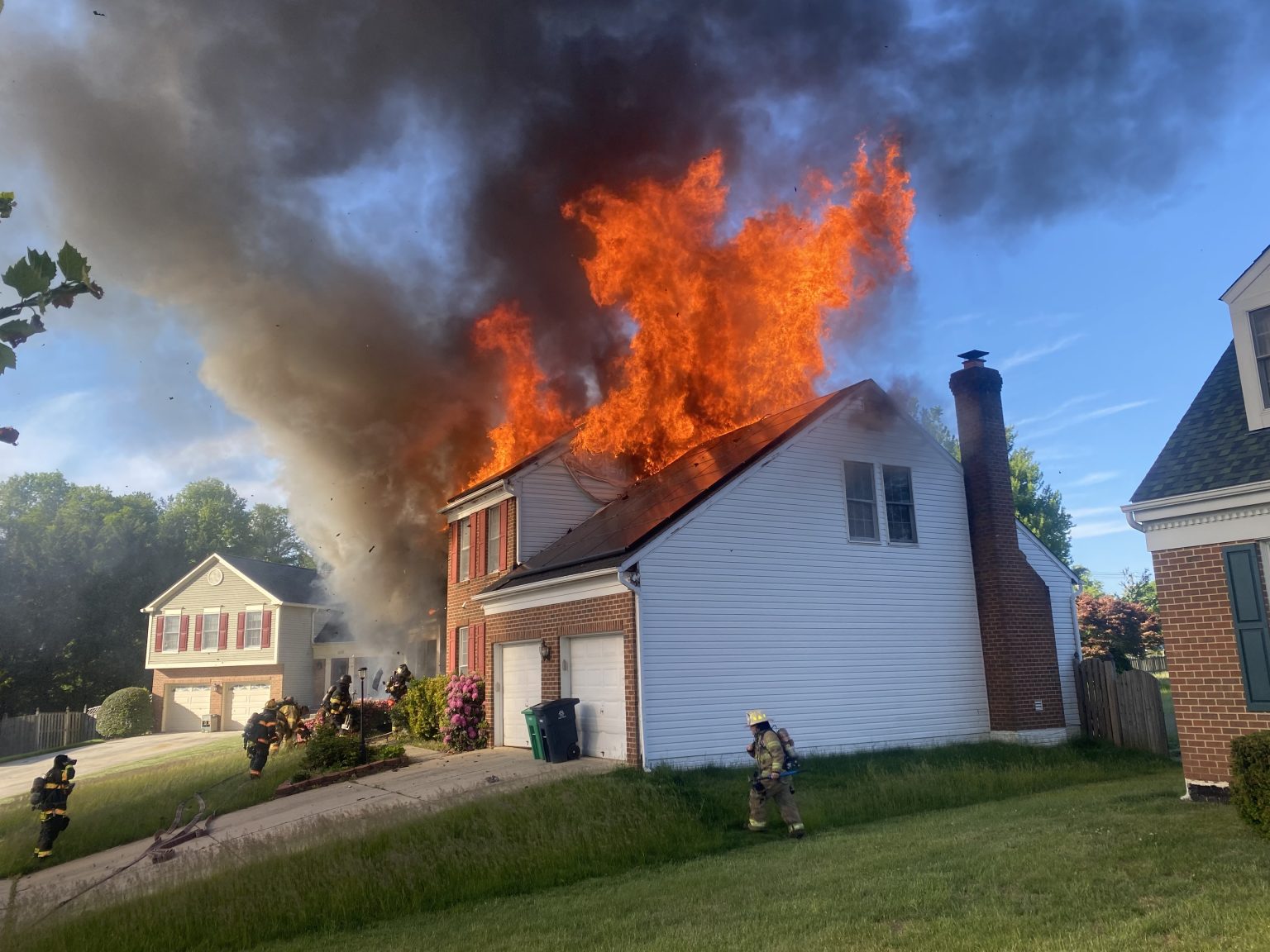 A house is engulfed in flames and thick black smoke, with fire mainly on the roof. Firefighters in protective gear are approaching the scene, and another house is visible nearby under a clear blue sky.