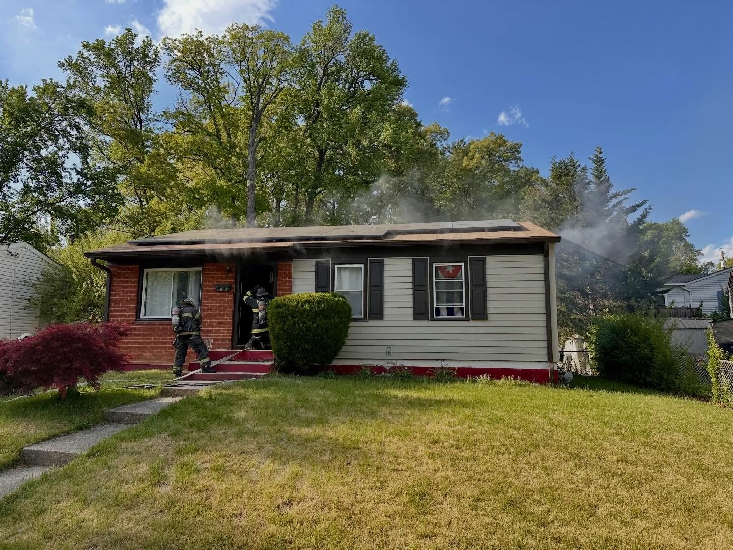 Two firefighters stand outside a single-story house with smoke coming from the roof. The house has beige siding, red brick, and black shutters, and is surrounded by green trees and grass under a blue sky.