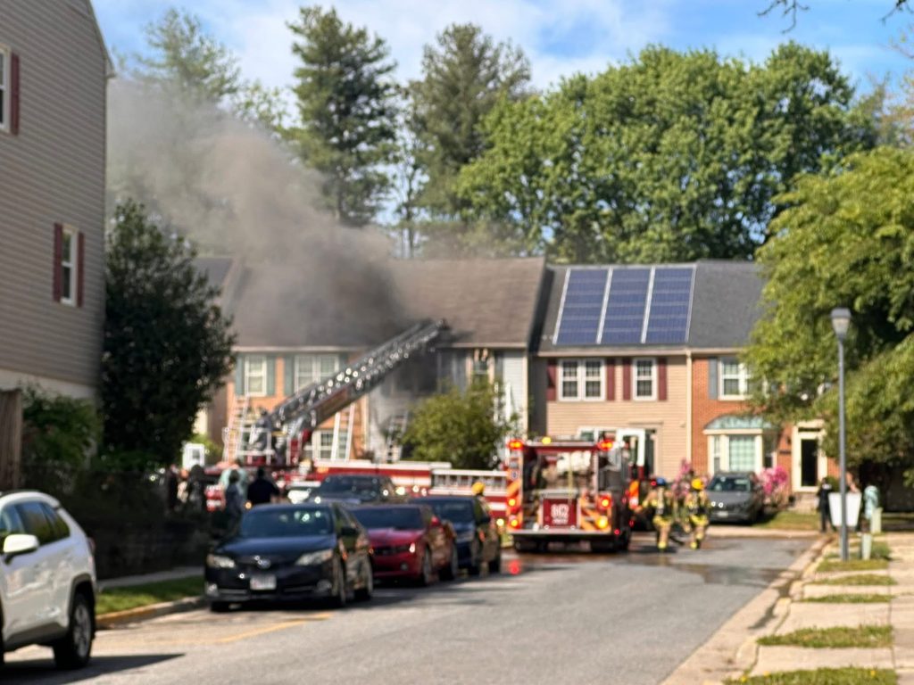 Firefighters respond to a house fire on a residential street, with smoke billowing from the roof. Fire trucks and emergency vehicles are present, and a ladder extends to the roof. Neighboring houses and parked cars are visible.