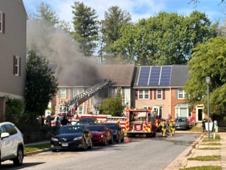 Firefighters respond to a house fire on a residential street, with smoke billowing from the roof. Fire trucks and emergency vehicles are present, and a ladder extends to the roof. Neighboring houses and parked cars are visible.