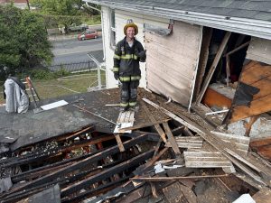 A firefighter in full gear stands on a damaged roof next to a house with a large hole and burned, broken wooden beams, showing the aftermath of a fire. Debris is scattered around.