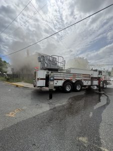 A fire truck is parked on a street while smoke billows from a building nearby. The sky is cloudy, and water is visible on the ground near the truck. Emergency response is underway.