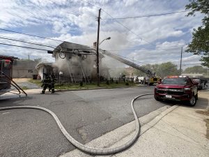 Firefighters work to extinguish heavy smoke coming from a house. Fire trucks, ladders, and hoses are in use, blocking a residential street under a partly cloudy sky. Emergency vehicles are present.