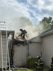 A firefighter in full gear stands on the roof of a house using an axe amid thick smoke, while two firefighters watch from below. A ladder leans against the building, and part of the home is damaged by fire.
