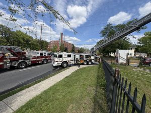 Several fire trucks are parked on a street near a building. A long ladder extends from one truck across the street. Trees, a fence, and a grassy area are in the foreground under a partly cloudy sky.