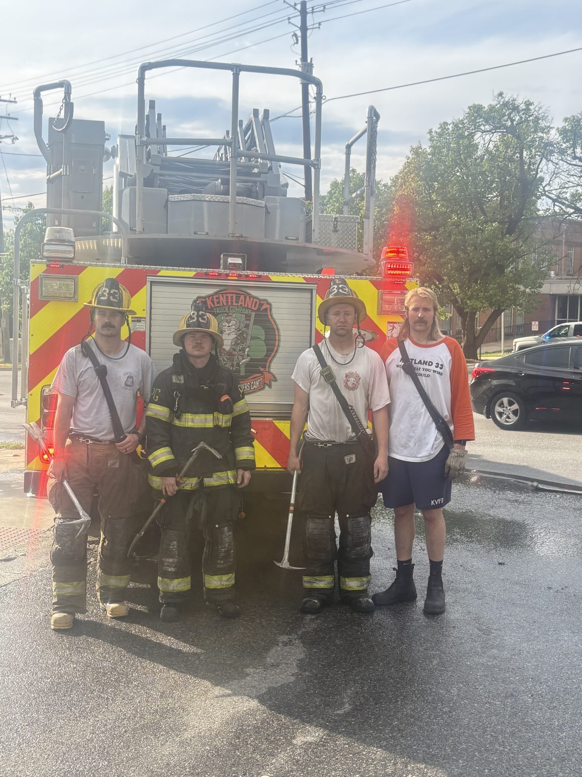 Four firefighters in gear stand in front of a fire truck labeled "Kentland Volunteer Fire Department" on a sunny day. The truck's lights are on, and a tree and buildings are visible in the background.