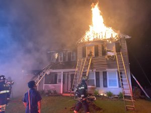 A two-story house is engulfed in flames on the roof at night. Smoke fills the air as firefighters in gear stand in the yard and ladders lean against the house, working to control the blaze.