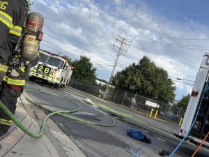 Firefighters stand by as a fire truck labeled "28" is parked on a street with hoses laid out on the ground. Trees, power lines, and a fenced area are visible under a partly cloudy sky.