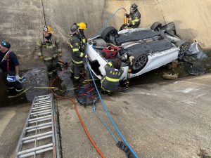 Firefighters work to rescue occupants from an upside-down white car after a crash in a concrete ditch. Emergency equipment, hoses, and a ladder are visible at the scene.