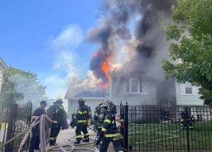 Firefighters in full gear respond to a house fire with heavy smoke and flames pouring from the roof. They prepare hoses near the burning two-story home while smoke billows into the blue sky.