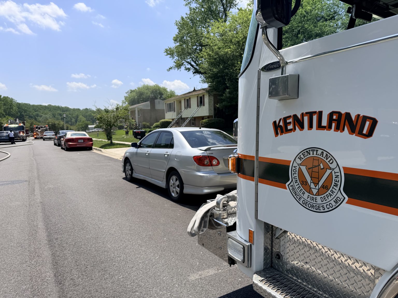 A Kentland fire truck is parked on a suburban street, with several cars and houses visible on a sunny day. Trees line the neighborhood, and the sky is clear with a few clouds.