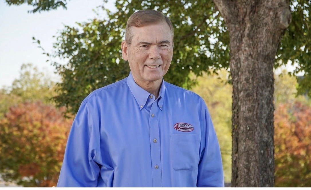 A man wearing a blue button-down shirt with a logo stands outdoors near a tree, smiling at the camera. The background features trees with autumn foliage.