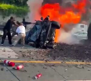 Three people try to help at the scene of a car crash where a vehicle is overturned and engulfed in flames, with fire extinguishers scattered on the road.